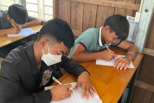 students sitting at wooden desks, writing on paper, with one student wearing a face mask