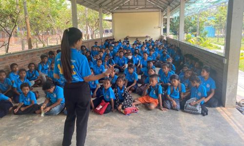 group of children sitting on the ground, listening to a woman speaking in front