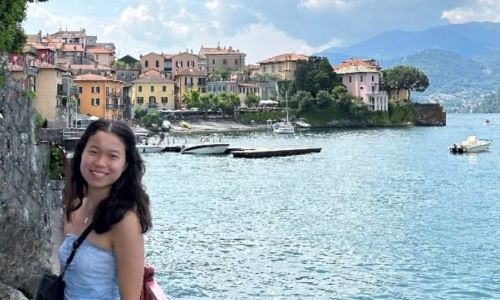 Smiling woman standing by a lakeside walkway with colorful buildings behind her