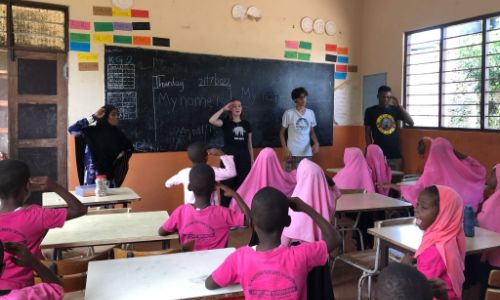 A group of teachers leads a classroom activity with students, practicing English greetings in front of a chalkboard.