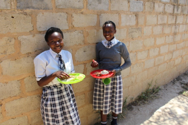 Two students in school uniforms enjoy lunch together while leaning against a brick wall.