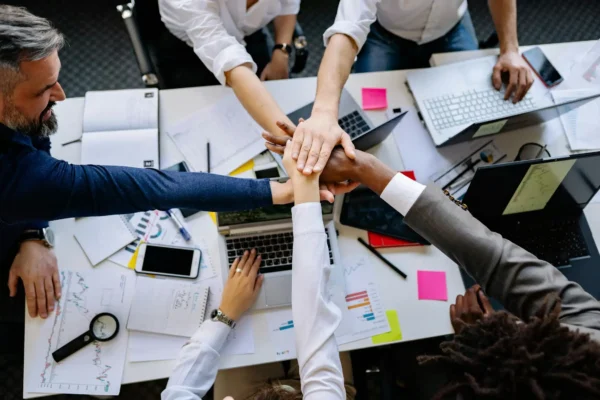 A team of interns join hands in a symbol of teamwork over a desk covered with laptops, papers, and office supplies.