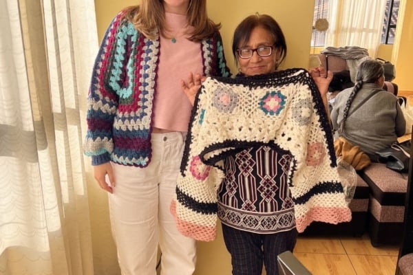 An intern from the Social Business Internship in Latin America posing with a local while holding up a knitted sweater.