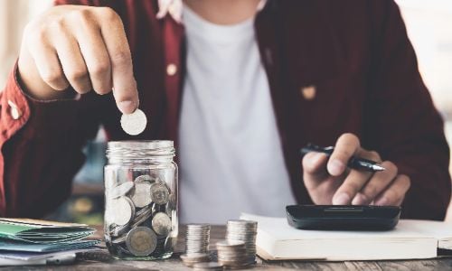 a man's hand putting money in the glass jar while using a calculator