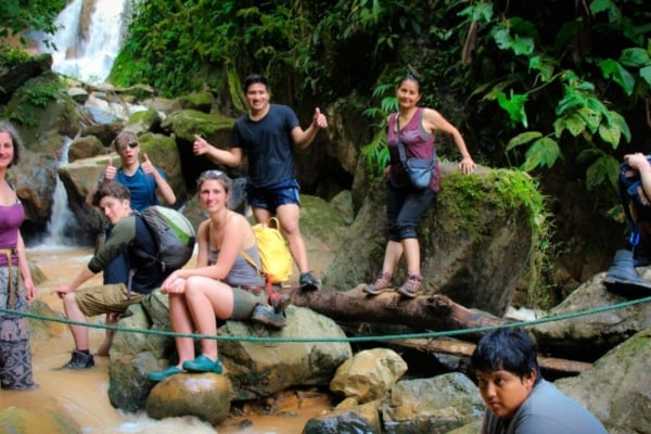 Group of people standing and sitting on rocks near a small waterfall in a forested area