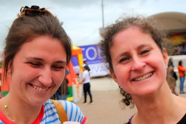 Two people smiling while standing outdoors, one holding a small cup, with event tents and people in the background