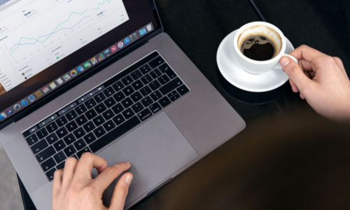 Remote worker using a laptop with data dashboards open while holding a cup of coffee