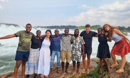 Nine people with their arms around one another standing on a cliff in front of some waves.